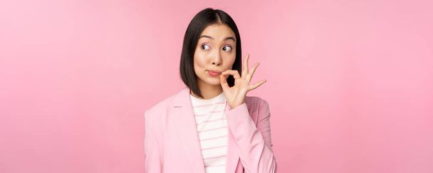 Portrait of asian corporate woman showing mouth seal, close shut lips on key gesture, promise keep secret, standing over pink background in suit - Photo, Image