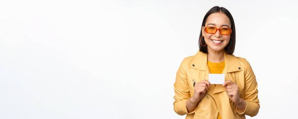 Портрет красивої сучасної азіатки в сонцезахисних окулярах, усміхнений щасливий, показує кредитну картку, що стоїть на білому тлі
 - Фото, зображення