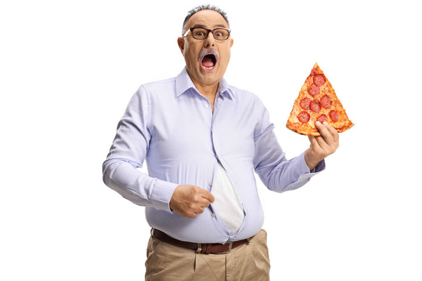 Shocked mature man in a tight shirt holding a slice of pepperoni pizza isolated on white background - Фото, изображение
