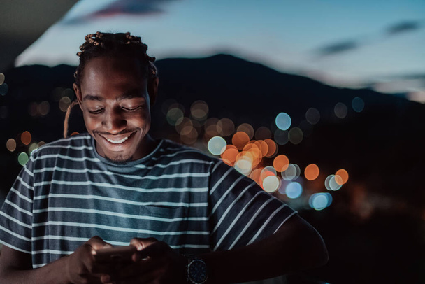 Ο νεαρός άνδρας σε ένα αστικό δρόμο της πόλης τη νύχτα γραπτών μηνυμάτων σε smartphone με bokeh και νέον φώτα της πόλης στο παρασκήνιο.  - Φωτογραφία, εικόνα