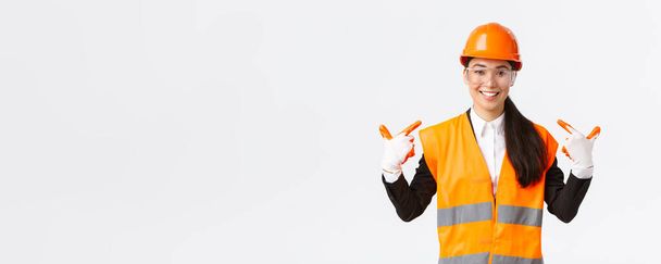 Smiling confident asian female chief construction engineer in safety helmet, gloves and glasses pointing at herself, notify to follow safe protocol and wearing special clothing, white background - Фото, зображення