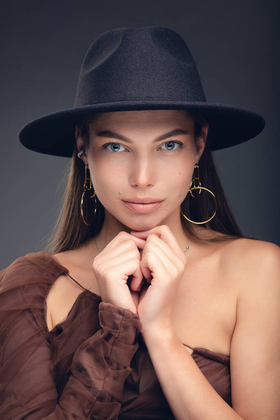 Fashionable studio portrait of a beautiful young girl in black hat on a grey background. - Фото, изображение