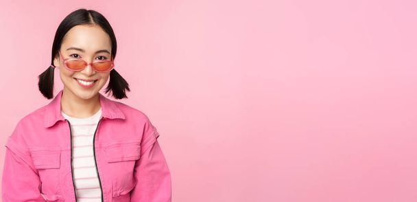 Gros plan de beau modèle féminin asiatique dans des lunettes de soleil élégantes, posant sur fond rose dans une tenue tendance, espace de copie - Photo, image