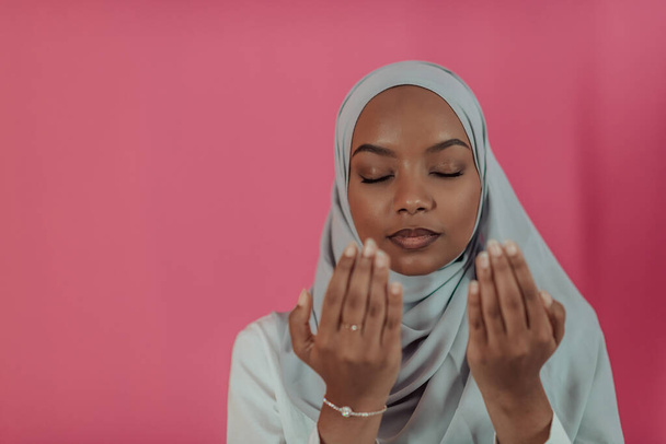 Modern African Muslim woman makes traditional prayer to God, keeps hands in praying gesture, wears traditional white clothes, has serious facial expression, isolated over plastic pink background - Photo, image