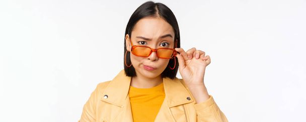 Genç Asyalı stil sahibi bir kadın tasviri, güneş gözlüklerinin altından kuşkucu, inançsız bir yüz ifadesiyle bakıyor, şüphe uyandırıyor, beyaz arka planda duruyor. - Fotoğraf, Görsel