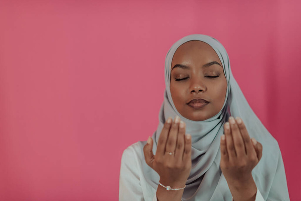 Modern African Muslim woman makes traditional prayer to God, keeps hands in praying gesture, wears traditional white clothes, has serious facial expression, isolated over plastic pink background - Fotografie, Obrázek