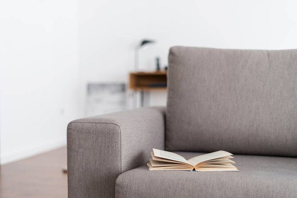 Zeit zum Lesen. Selektiver Fokus auf aufgeschlagenes Papierbuch in gebundenem Einband auf grauem Sofa liegend. Nahaufnahme von ergonomischem Grau in komfortablem Haus. Erholungs-, Entspannungskonzept - Foto, Bild