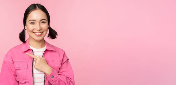 Portret pięknej uśmiechniętej Azjatki, z lewej strony palca, pokazujący reklamę, baner promo, stojący nad różowym tłem - Zdjęcie, obraz