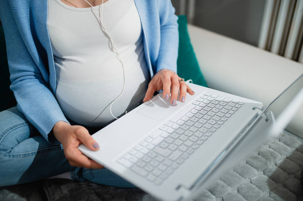 白人の妊婦はソファに座り、白いラップトップとイヤホンを使用します。彼女の顔は見えない。右手でノートパソコンを持ち、左手でタイプする。女は足を組んで座る. - 写真・画像