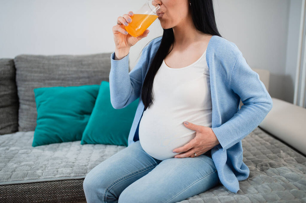 Egy terhes nő narancslevet iszik a nappaliban, és a bal kezével fogja a hatalmas hasát. Leül a kanapéra, és élvezi a szükséges vitaminokat. Zöld párnák vannak a háttérben..  - Fotó, kép