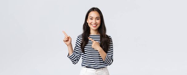 Lifestyle, people emotions and casual concept. Good-looking young korean female advertise product, smiling camera excited and pointing fingers upper left corner at promo banner - Photo, Image
