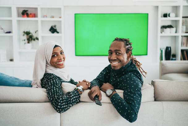 African Couple Sitting On Sofa Watching TV Together Chroma Green Screen Woman Wearing Islamic Hijab Clothes - Fotoğraf, Görsel