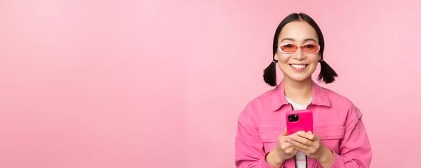 Портрет корейской девушки в стильных солнцезащитных очках с мобильным телефоном, с помощью приложения для смартфона, стоящей на розовом фоне - Фото, изображение