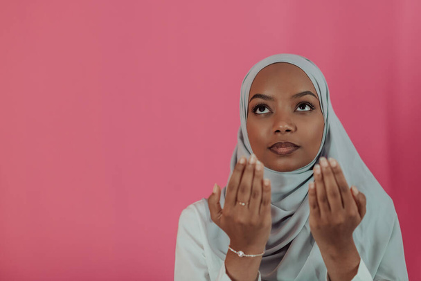 Modern African Muslim woman makes traditional prayer to God, keeps hands in praying gesture, wears traditional white clothes, has serious facial expression, isolated over plastic pink background - Zdjęcie, obraz