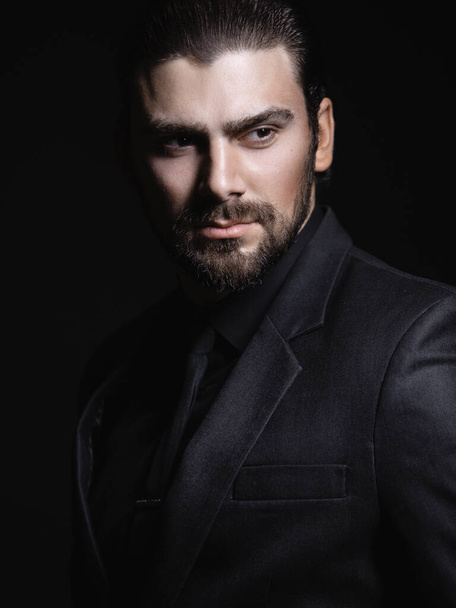 Young white Caucasian man on a black background in a black jacket, shirt and black tie looks out from under his forehead. Strict male portrait. The guy in the classic black suit. - Photo, Image