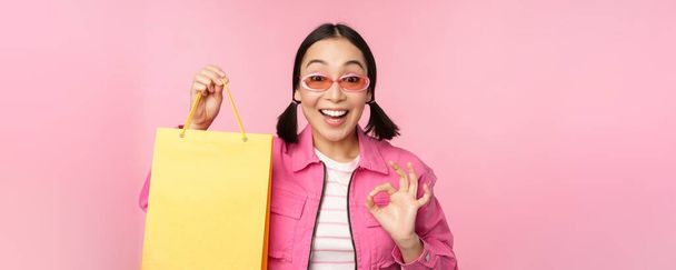 Shopping. Stylish asian girl in sunglasses, showing bag from shop and smiling, recommending sale promo in store, standing over pink background - Photo, Image