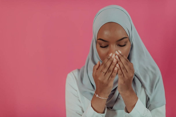 Moderna mujer musulmana africana hace la oración tradicional a Dios, mantiene las manos en el gesto de oración, lleva ropa blanca tradicional, tiene una expresión facial seria, aislado sobre fondo rosa plástico - Foto, imagen