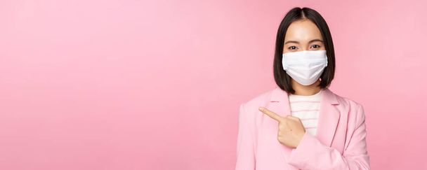 Portret azjatyckiej bizneswoman w medycznej masce i garniturze, wskazując palcem w lewo, pokazując reklamę, firmowy baner, studio różowe tło - Zdjęcie, obraz