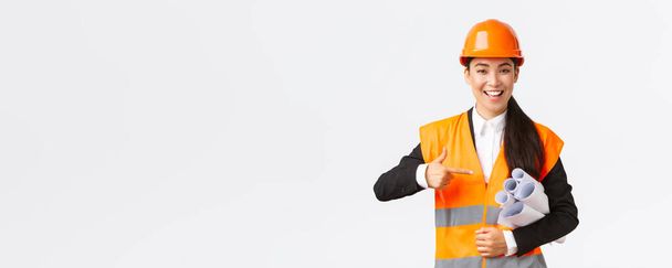 Professional confident asian female architect, chief engineer in safety helmet pointing finger at blueprints, showing project plan or documents for construction, standing white background - Photo, Image