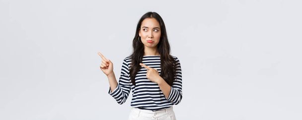 Lifestyle, beauty and fashion, people emotions concept. Disappointed sad asian girl complaining, feeling regret or jealousy as pouting uneasy and pointing fingers left at promo banner - Photo, Image
