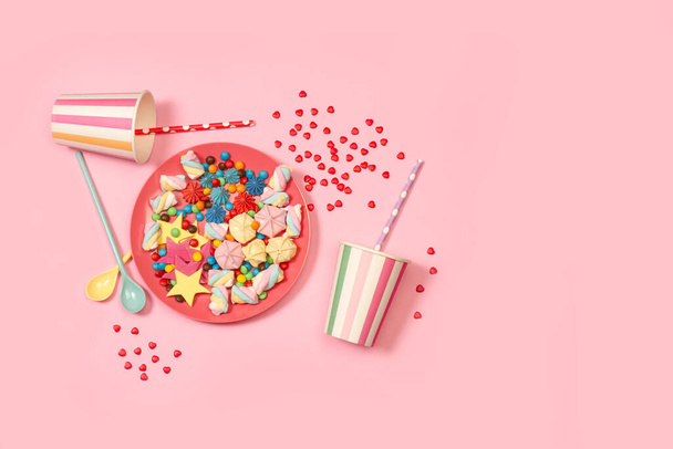Meringues et bonbons colorés dans une assiette rose et des tasses jetables sur un fond rose - Photo, image