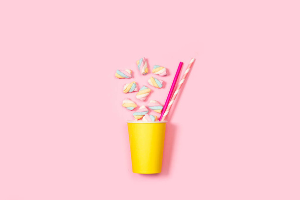 Drinking straws and marshmallows in a yellow paper glass on a pink background - Фото, изображение