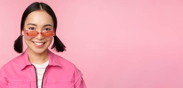 Közel a stílusos koreai lány napszemüvegben, mosolyogva boldog, pózol a rózsaszín háttér. Az emberek szembesülnek - Fotó, kép