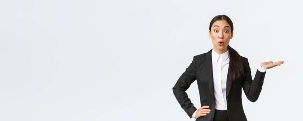 Verbazingwekkende Aziatische zakenvrouw in zwart pak wijzend hand rechts en kijk naar camera geïnteresseerd, bespreken aankondiging, tonen banner, staande witte achtergrond geïntrigeerd - Foto, afbeelding