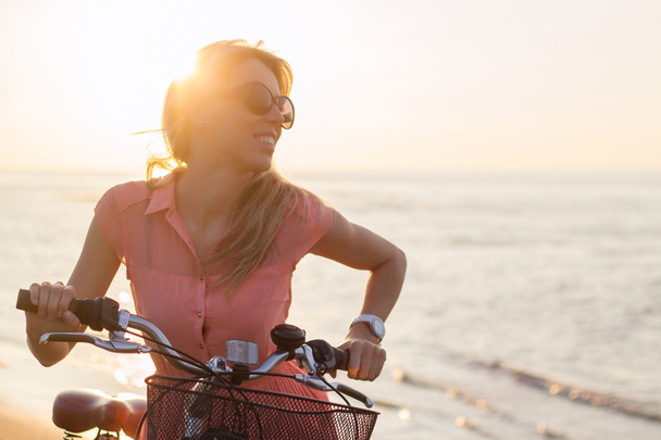 Young fashionable woman riding bicycle on the beach at sunset - Photo, image