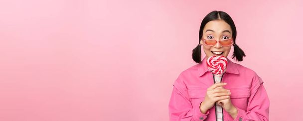 Глупая и милая азиатская модель лижет леденец, ест сладкие конфеты и улыбается, выглядит взволнованным, стоя на розовом фоне - Фото, изображение