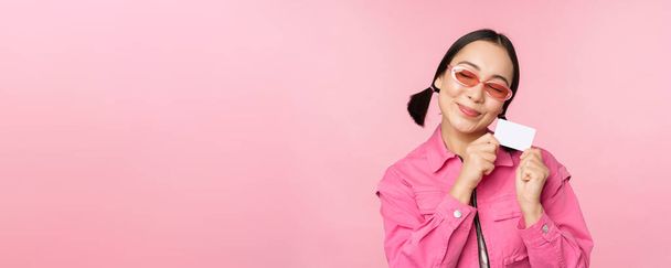 Porträt eines stilvollen, modernen asiatischen Mädchens, zeigt Rabatt, Kreditkarte und sieht zufrieden aus, zahlt kontaktlos, Konzept des Einkaufs, steht über rosa Hintergrund - Foto, Bild