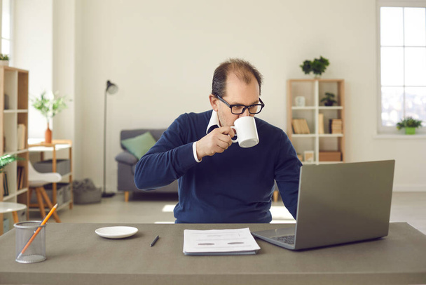 Mature caucasian man in eyeglasses working on laptop drinking coffee at home - 写真・画像