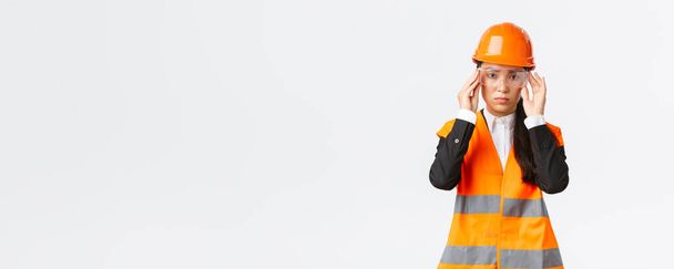 Серьёзно выглядящая молодая азиатка-строительный менеджер следует протоколу безопасности, носит защитные очки и шлем перед входом на предприятие, стоя на белом фоне - Фото, изображение