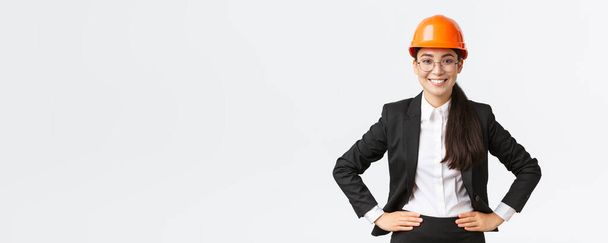 Profesjonalny uśmiechnięty azjatycki inżynier budowlany, architekt w czarnym garniturze i kasku bezpieczeństwa wygląda zadowolony w pracy, stoi zadowolony, businesswoman sprawdza wszystko na placu budowy - Zdjęcie, obraz