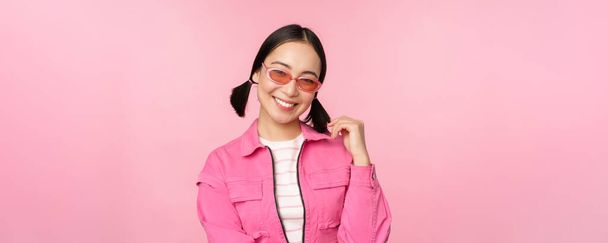Mooi aziatisch meisje in zonnebril, glimlachen op camera, poseren tegen roze studio achtergrond - Foto, afbeelding