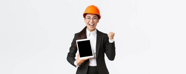 Successful winning female engineer at factory, construction manager in safety helmet and business suit fist pump, triumphing over achievement, showing digital tablet display, graph or diagram - Zdjęcie, obraz