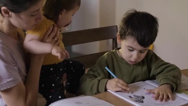 Vorschulkind sitzt am Tisch und malt Bild in Album, kaukasische Mutter und kleine Tochter sitzen neben dem Jungen. Schöne junge Mädchen - Filmmaterial, Video
