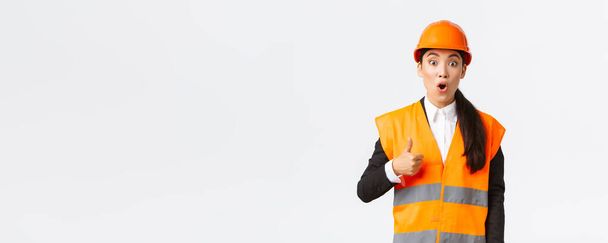 Impressed and excited asian female engineer in safety helmet and reflective jacket fascinated with nice work, showing thumbs-up gasping astounded, say well done, great job, standing white background - Foto, Imagen
