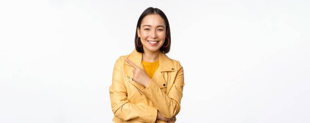 Портрет усміхненої азіатки, що вказує пальцем ліворуч, показує логотип або простір для копіювання, рекламу на порожній стороні, що стоїть на білому тлі
 - Фото, зображення