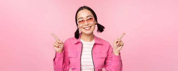 Elección. Elegante chica coreana, modelo femenino asiático señala los dedos de lado, muestra dos variantes, anuncio de producto, elementos de demostración, de pie sobre fondo rosa - Foto, imagen