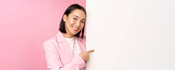 Portret szczęśliwej uśmiechniętej japońskiej korporacyjnej kobiety wskazującej palcem na pokładzie, pokazującej smth na pustej przestrzeni, stojącej w garniturze na różowym tle - Zdjęcie, obraz