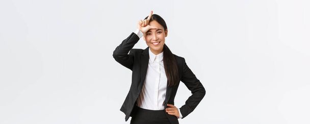 Успешная азиатская бизнесвумен в черном костюме высмеивает своих соперников, показывает знак проигравшего как победу и празднование победы над конкурентами, стоя довольной на белом фоне - Фото, изображение