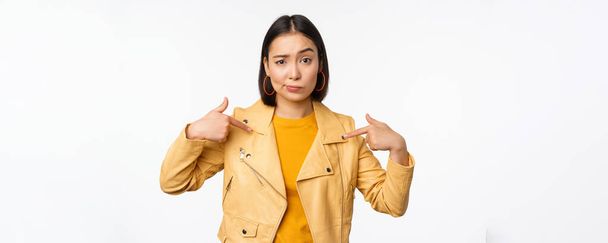 Image of unamused asian girl pointing at herself, looking with disbelief skeptical at camera, posing against white background - Photo, Image