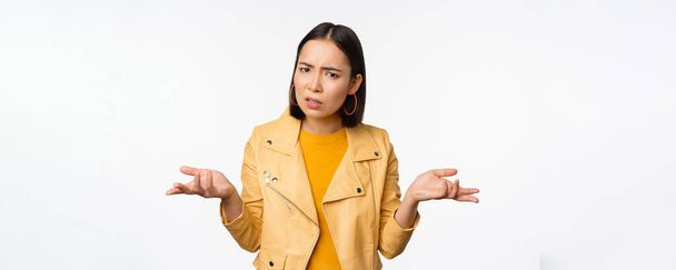 Image of confused young asian woman shrugging shoulders, looking puzzled and clueless at camera, cant understand, standing against white background - Photo, Image