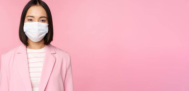Portrait of asian businesswoman in medical face mask, wearing suit, concept of office work during covid-19 pandemic, standing over pink background - Photo, image