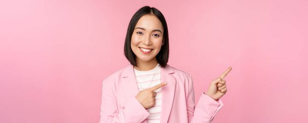 Enthousiaste professionele zakenvrouw, verkoopster wijst vingers naar rechts, toont reclame of bedrijfslogo opzij, poseren over roze achtergrond - Foto, afbeelding