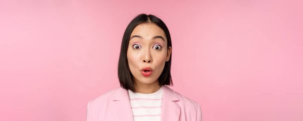 De cerca retrato de mujer corporativa asiática, mujer de negocios mirando sorprendido y sorprendido en la cámara, de pie en traje sobre fondo rosa - Foto, Imagen