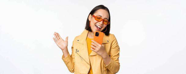 Conceito de tecnologia e pessoas. Feliz dançando menina asiática em óculos de sol, usando smartphone, cantando e sorrindo, de pé sobre fundo branco - Foto, Imagem