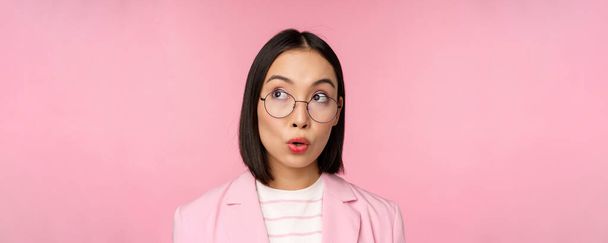 Entusiástica vendedora, asiática empresária de óculos, olhando intrigada no canto superior esquerdo logotipo, anúncio da empresa, de pé sobre fundo rosa - Foto, Imagem