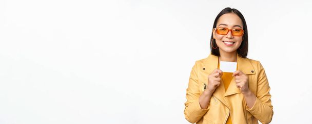 Πορτρέτο του χαμογελαστού κορεάτικου γυναικείου μοντέλου σε γυαλιά ηλίου, που δείχνει την πιστωτική κάρτα, στέκεται πάνω από λευκό φόντο - Φωτογραφία, εικόνα